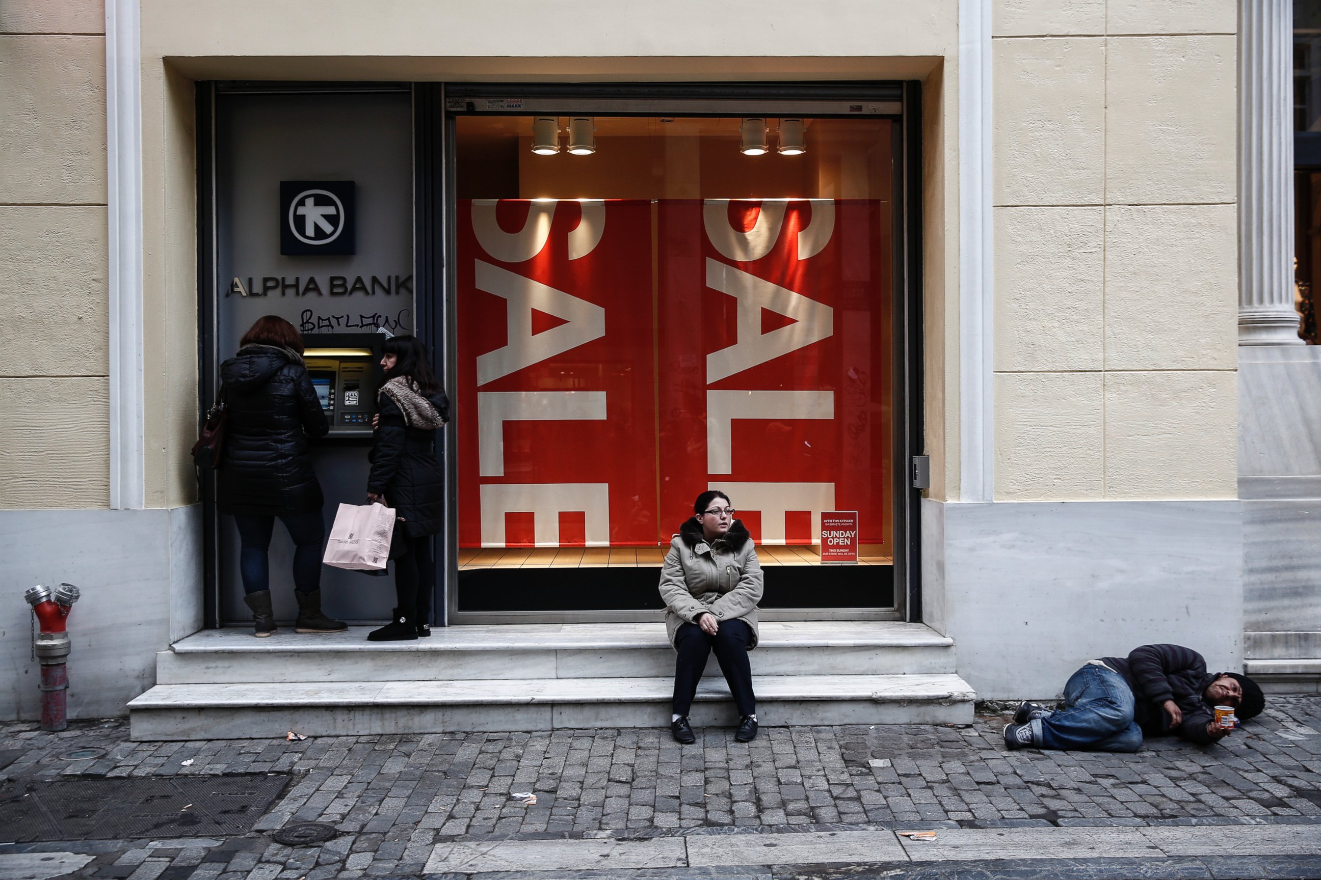 PHOTO: Women make a transaction at an ATM as a man begs on Ermou Street, Athens' main shopping strip