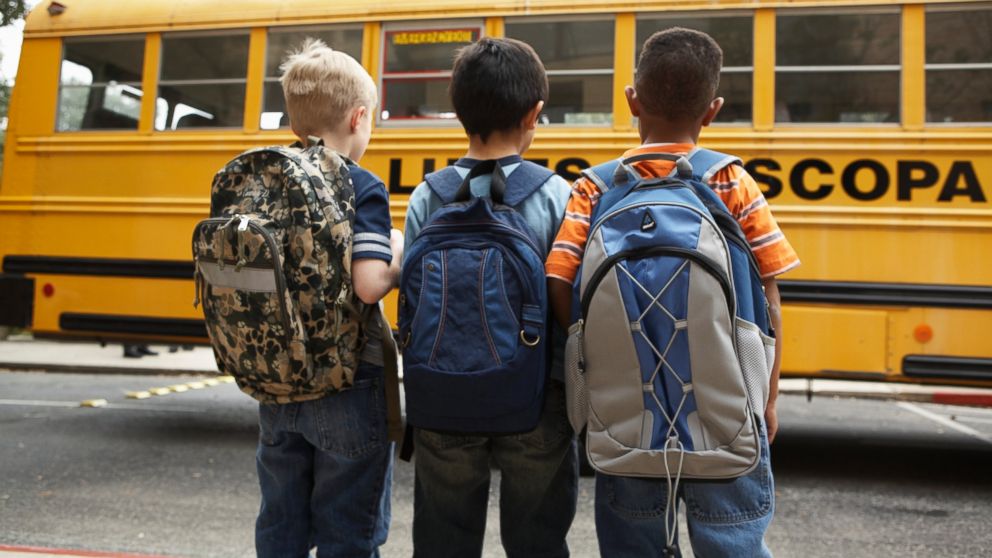 Three boys wearing backpacks stand near a school bus in this stock photo.