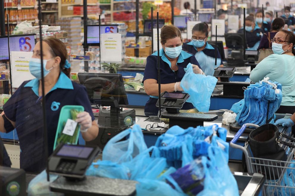 PHOTO: Cashiers stands behind a partial protective plastic screen and wear a mask and gloves as they work at the Presidente Supermarket on April 13, 2020, in Miami.