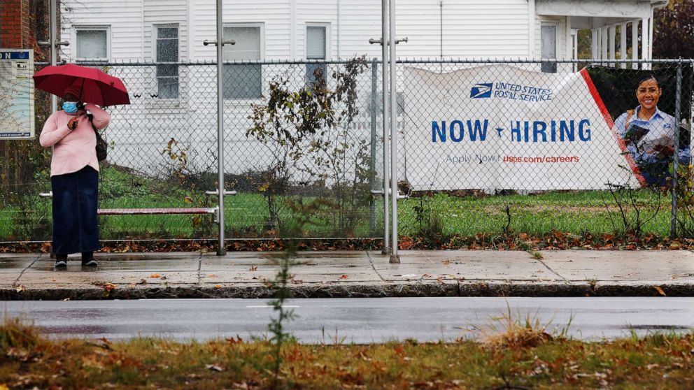 PHOTO: A woman waits for a bus next to a "Now Hiring" sign from the United States Postal Service in Boston, Oct. 30, 2021.