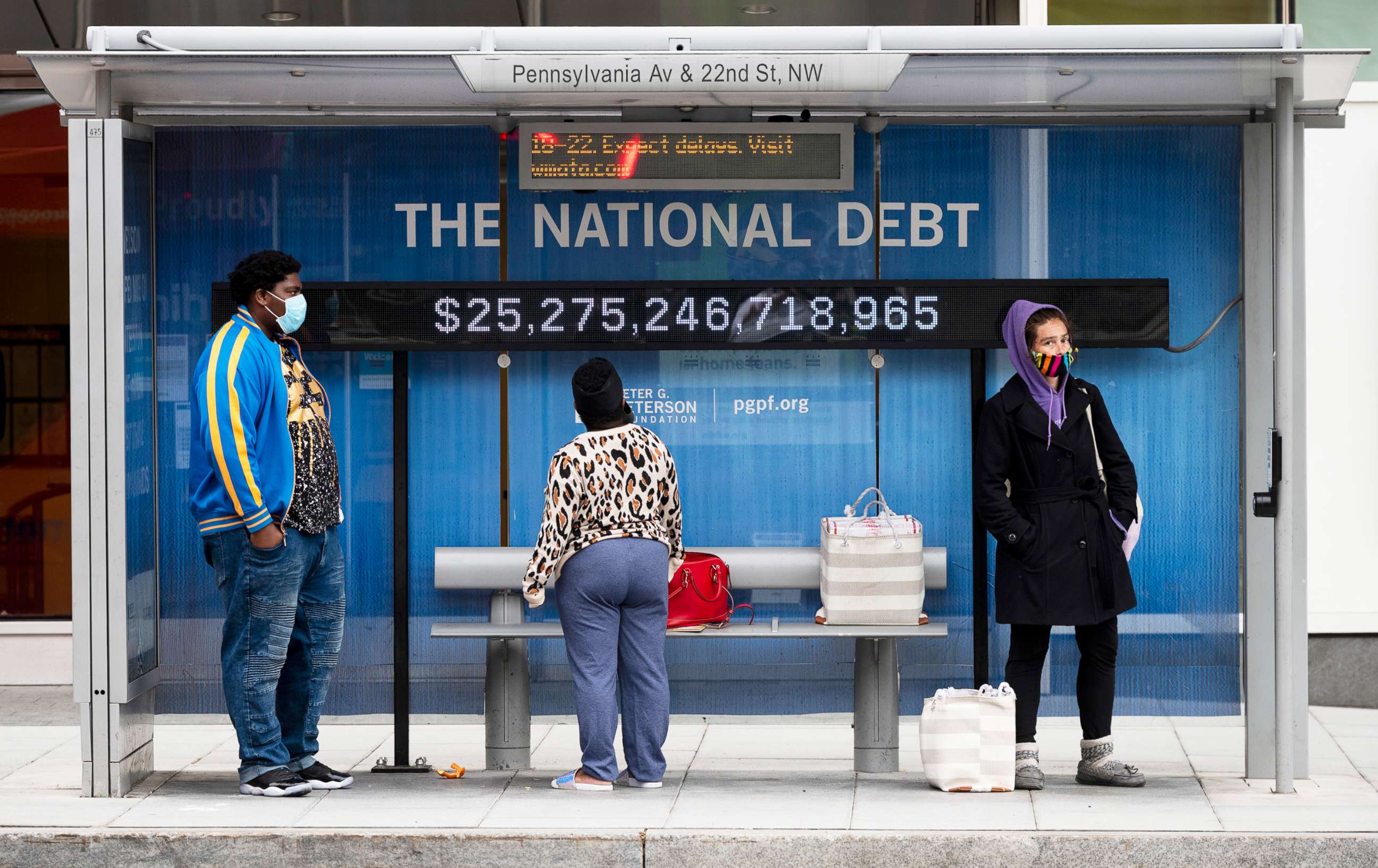 PHOTO: Passengers wearing face masks wait for their bus in front of a national debt display on Pennsylvania Ave. NW in Washington, May 18, 2020. 