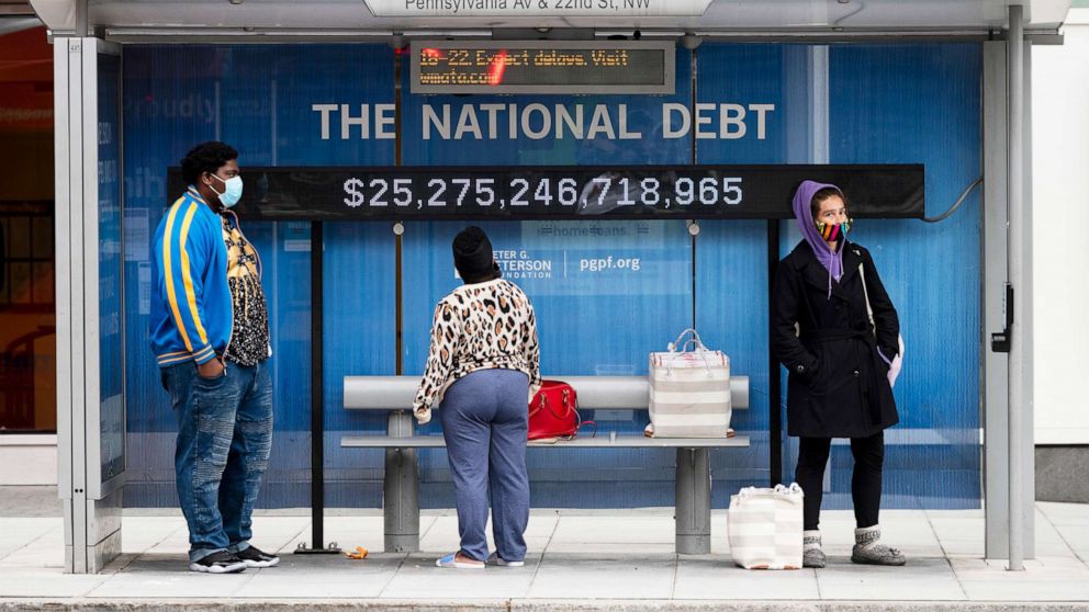 PHOTO: Passengers wearing face masks wait for their bus in front of a national debt display on Pennsylvania Ave. NW in Washington, May 18, 2020. 