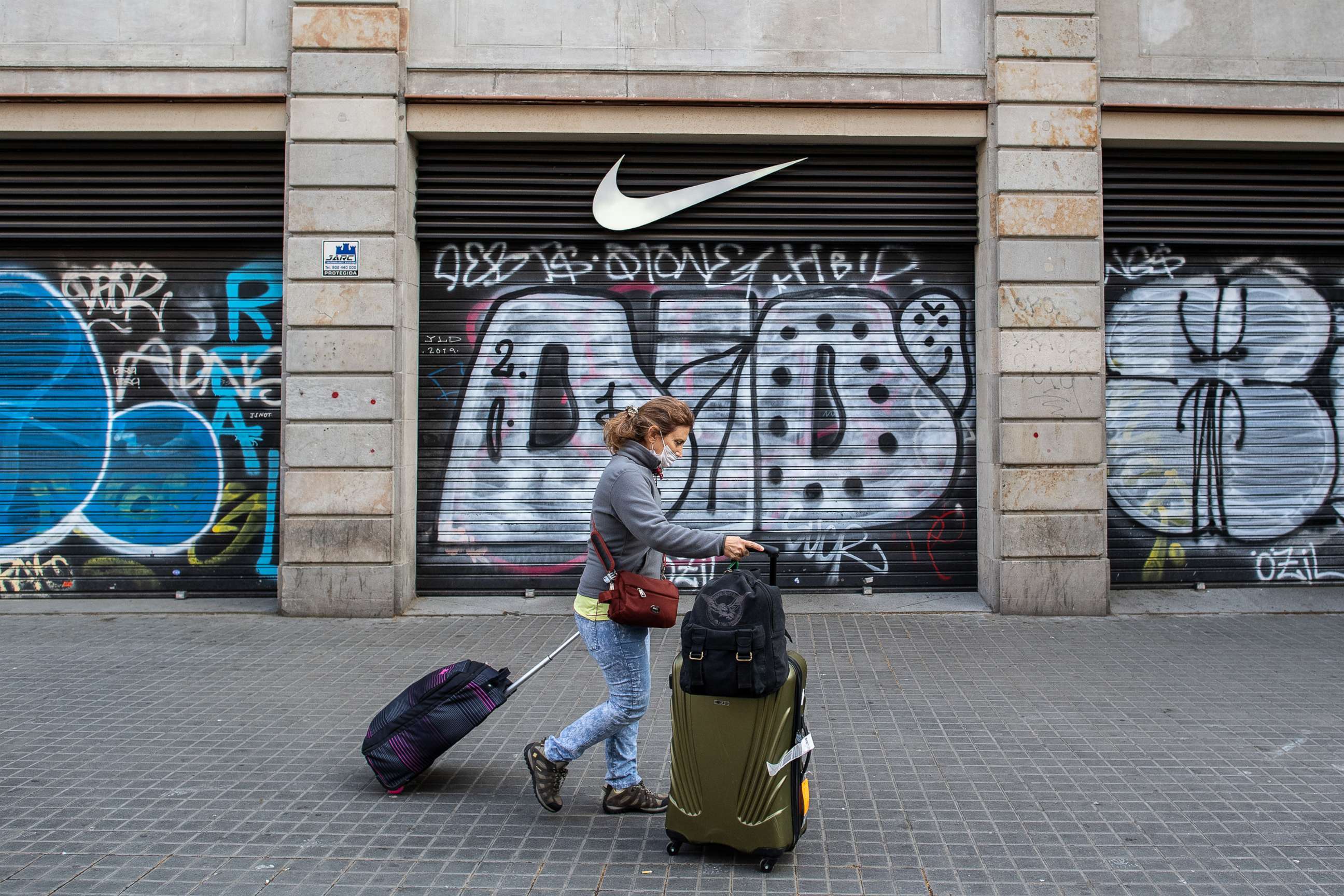 PHOTO: A tourist wears a protective mask as she carries her suitcases past a closed Nike store at Las Ramblas, March 15, 2020, in Barcelona, Spain. 