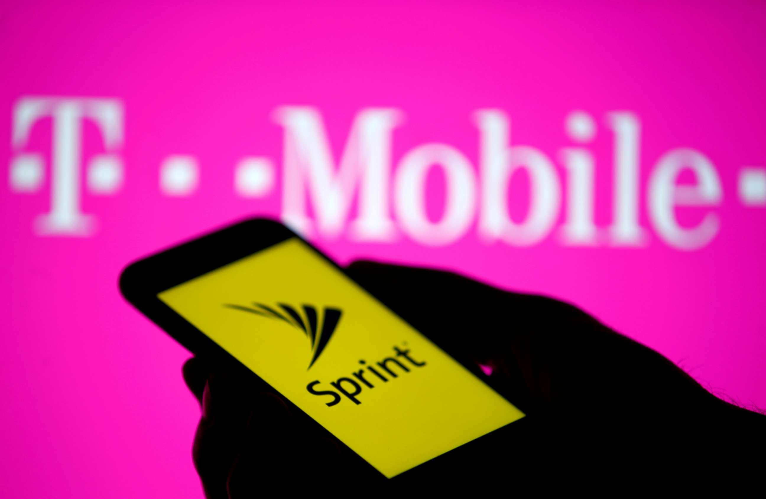 PHOTO: A posed photo shows a smartphone with Sprint logo in front of a screen projection of T-mobile logo.