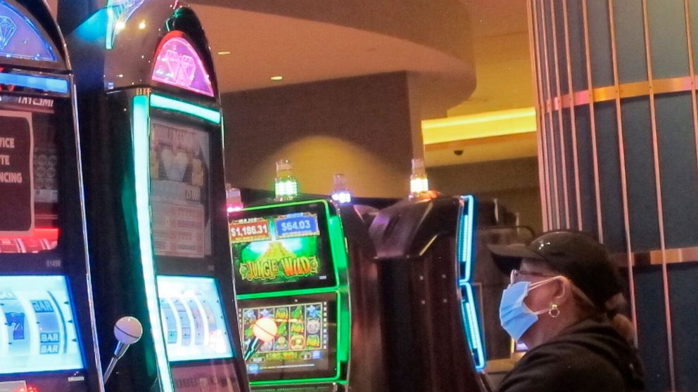 FILE - This May 3, 2021 file photo shows an intentionally disabled slot machine next to a woman playing a different slot machine while wearing a face mask at the Hard Rock casino in Atlantic City, N.J. Participants in a major casino conference Thursd