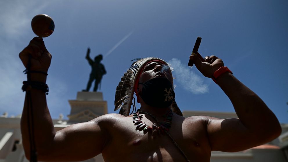 FILE - Gypsy Cordova blows smoke and shakes a maraca in front of a Juan Ponce de Leon monument while leading a group of activists in a march demanding statues and street names commemorating symbols of colonial oppression be removed, in San Juan, Puer
