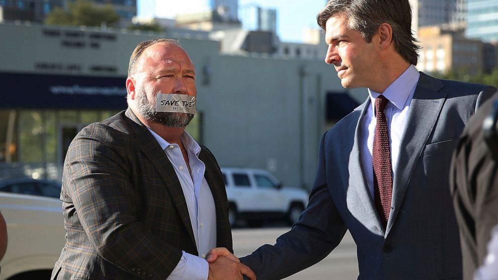 FILE - Alex Jones, left, arrives at the Travis County Courthouse in Austin, Texas, on July 26, 2022, with a piece of tape over his mouth that reads "Save the 1st." He shook hands with his lawyer, Andino Reynal. Although Jones portrays the lawsuit aga