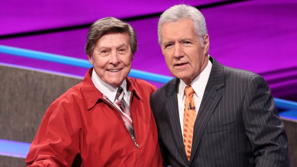 In this 2014 image provided by Jeopardy Productions, Inc., show announcer Johnny Gilbert, left, and game show host Alex Trebek appear on the set of "Jeopardy!" Gilbert worked with Trebek for 37 years and was reluctant to continue after losing his adm
