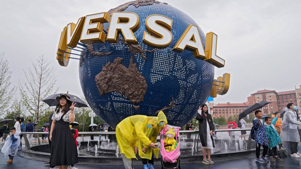 A family wearing raincoats and face masks to help protect themselves from the coronavirus pose for a selfie with an icon near the entrance to the Universal Studios Beijing in Beijing, Monday, Sept. 20, 2021. Thousands of people brave the rain visit t