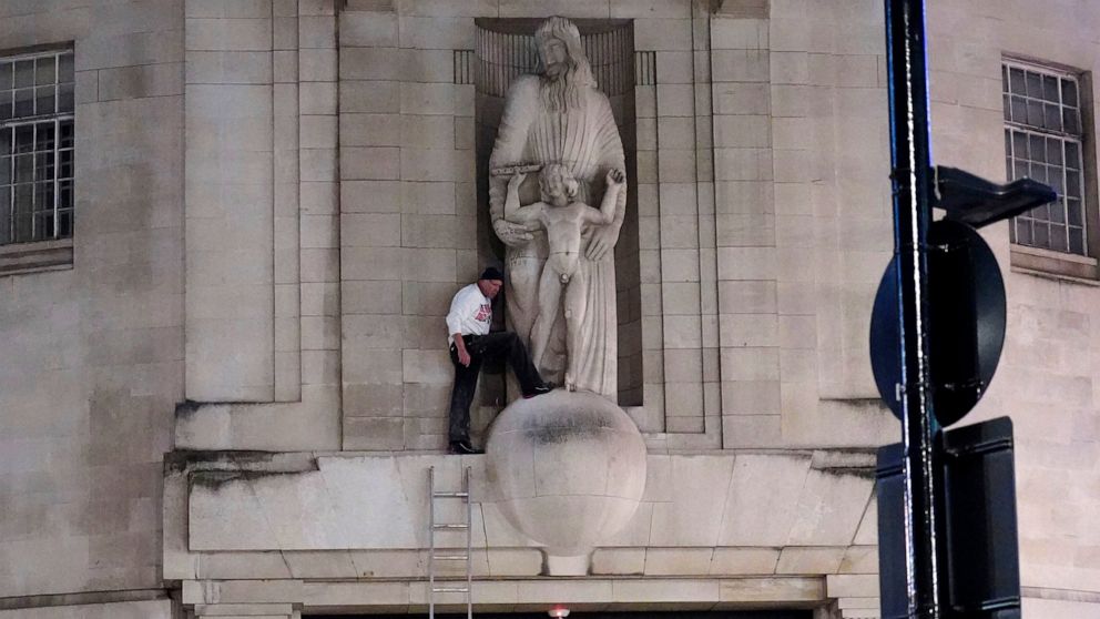 A man, after climbing a ladder, stands next to the statue of Prospero and Ariel from Shakespeare's play The Tempest by the sculptor Eric Gill outside the BBC's Broadcasting House in central London, Wednesday Jan. 12, 2022. British police have cordone