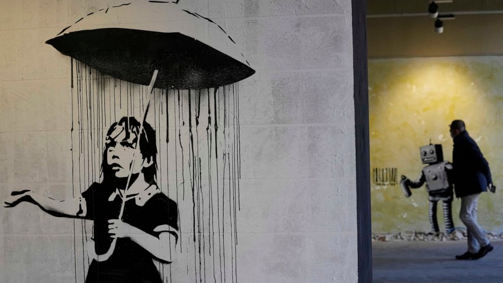 "The Umbrella girl" a reproduction of a mural by British artist Banksy is seen as a visitor walks past in background during the unveiling of the "The World of Banksy, The Immersive Experience" exhibition, in Milan, Italy, Thursday, Dec. 2, 2021. An e