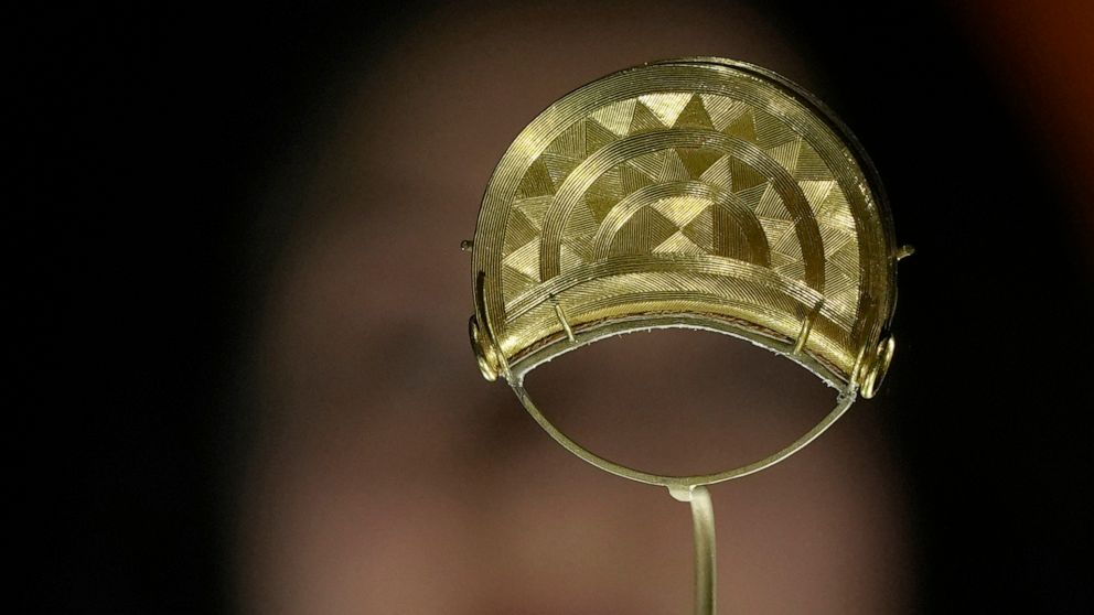 A member of staff poses next to a gold broach from Shropshire, England. 1000 BCE, on display at the The World of Stonehenge' exhibition at the British Museum in London, Monday, Feb. 14, 2022. The exhibition which displays objects and artifacts from t