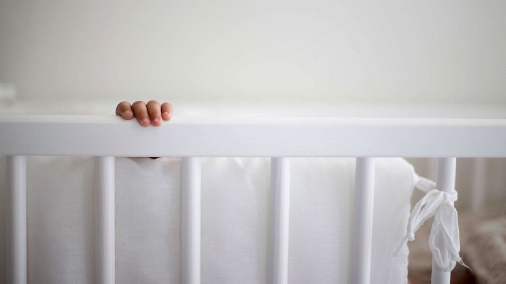 PHOTO: A baby's hand reaches for the rail of a crib that has a bumper tied to the rails.
