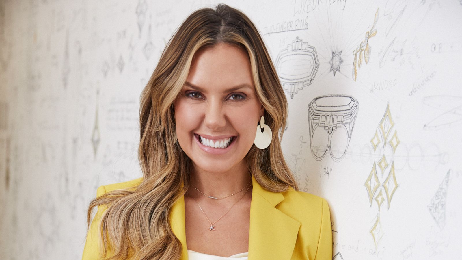 PHOTO: Fashion designer Kendra Scott wears a yellow blazer and blue jeans while leaning against white wall.