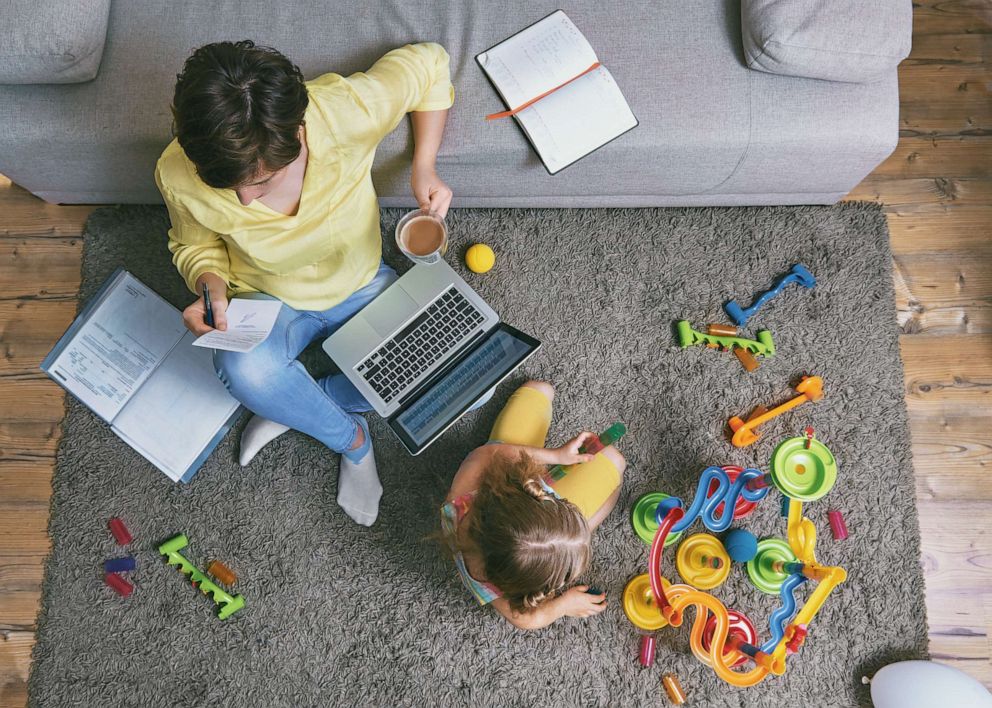 PHOTO: A woman works remotely from home while a child plays with blocks sitting on a carpet.