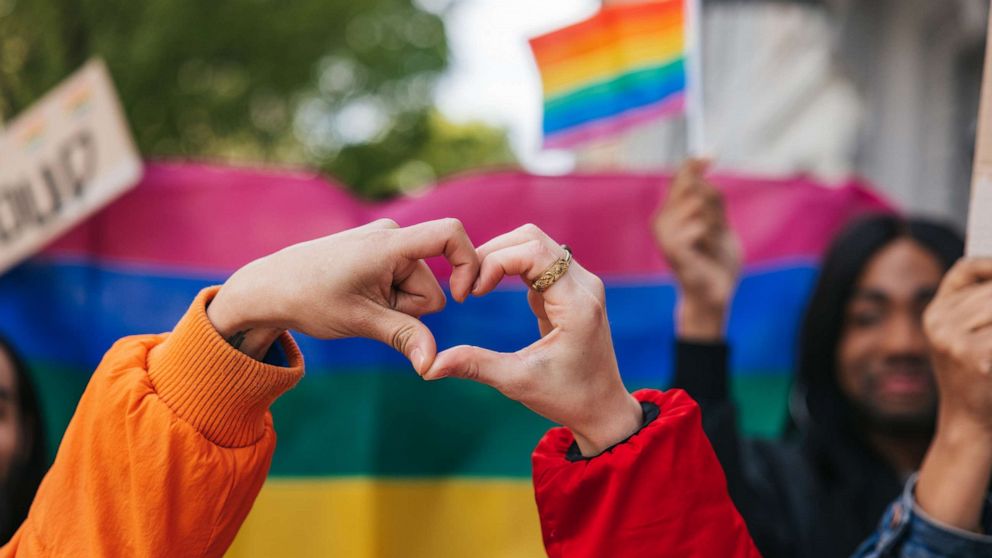 PHOTO: LGBTQIA+ friends making a heart shape against a rainbow flag celebrating love during a Pride Parade.