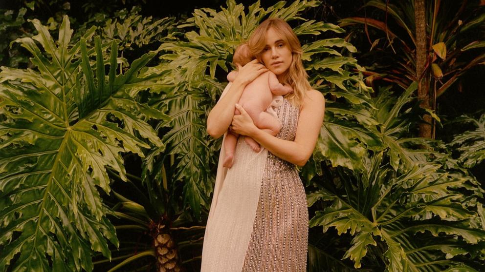 PHOTO: Image shows Suki Waterhouse standing on a lawn with lush green plants behind her, holding her baby daughter in her arms. The baby has her back to the camera, with her head facing over Suki’s shoulder. 