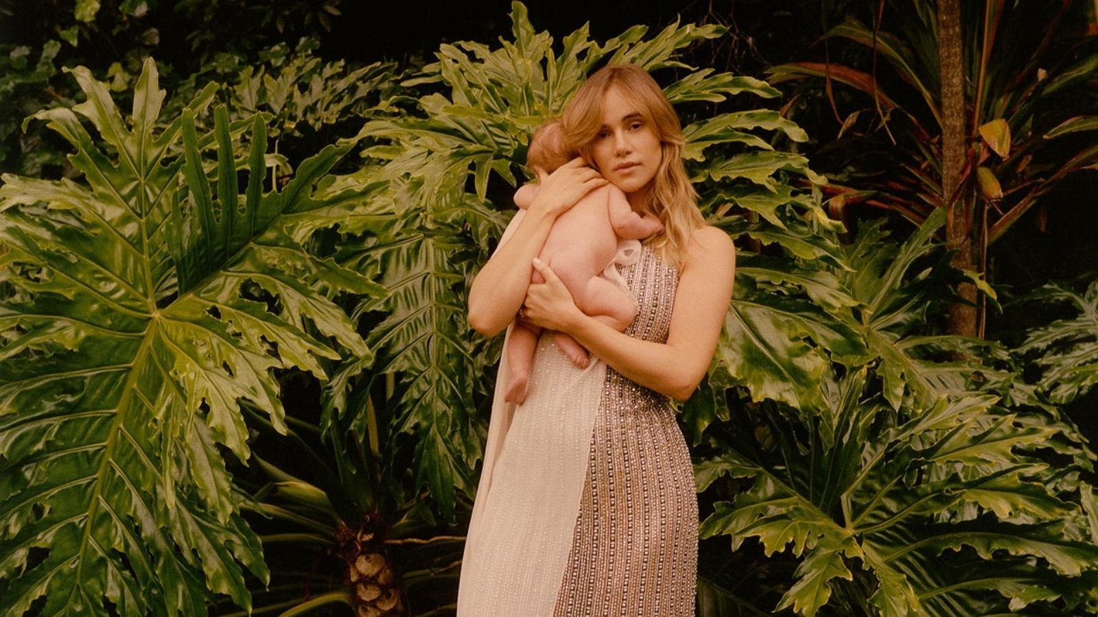 PHOTO: Image shows Suki Waterhouse standing on a lawn with lush green plants behind her, holding her baby daughter in her arms. The baby has her back to the camera, with her head facing over Suki’s shoulder.