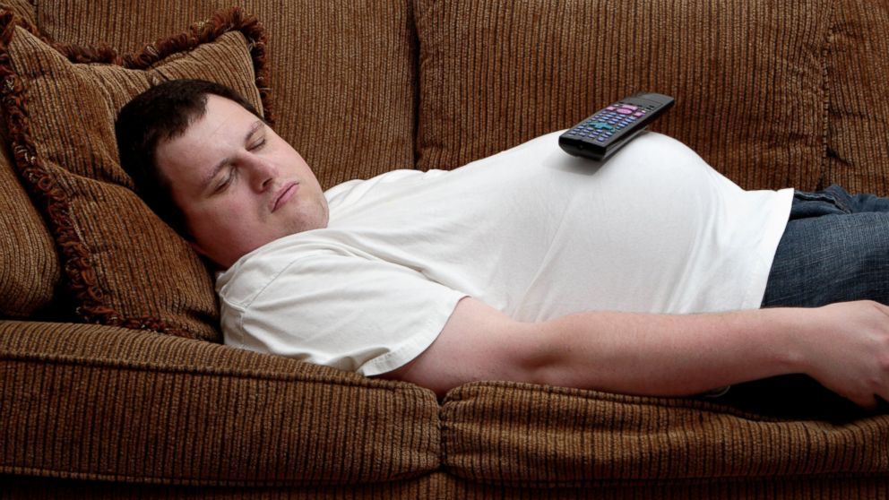 A man sleeps on the couch, with a TV remote control balancing on his protruding stomach.