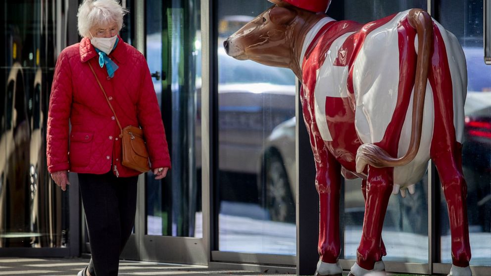 An elderly woman wearing a face mask walks past a plastic cow that decorates a hotel entrance in Frankfurt, Germany, Monday, March 29, 2021. (AP Photo/Michael Probst)
