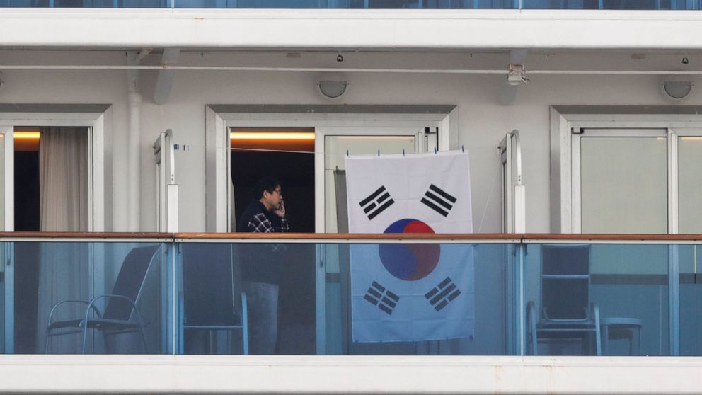 In this Feb. 15, 2020, photo, a man talks on the phone next to a South Korean flag hanging in the balcony of his cabin on the quarantined Diamond Princess cruise ship in Yokohama, near Tokyo. The 14-day quarantine for those on the ship is scheduled t