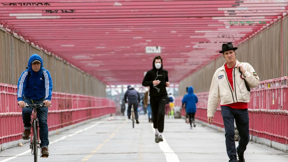 In this Monday, May 11, 2020 photo, a jogger wearing a face masks runs in between a biker and a pedestrian not wearing masks as they make their way over the Williamsburg bridge in New York.New York's governor has ordered masks for anyone out in publi