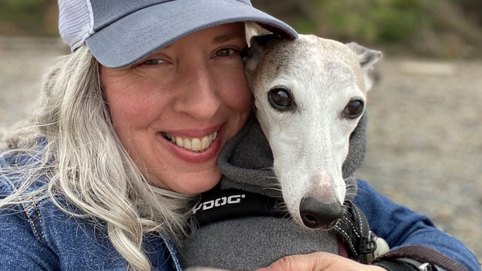 This photo provided by Amy Watson of Portland, Ore., shows her with her dog during a trip to the Oregon Coast in June 2022. Watson, approaching 50, says she has “never had any kind of recovery” from COVID-19. She has had severe migraines, plus digest