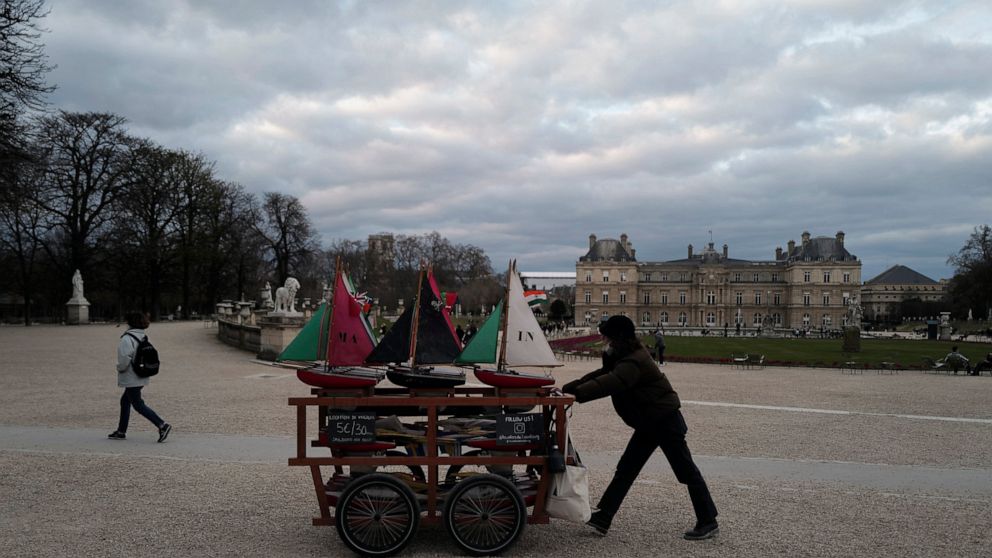 A woman pushes a cart of little sailing boats in the Luxembourg Garden with the Senat in background , in Paris, Sunday, March 21, 2021. The French government has backed off from ordering a tough lockdown for Paris and several other regions despite an