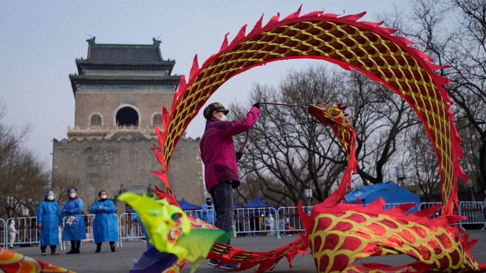 Community workers wearing protective suits watch over a masked resident twirling a dragon shaped ribbon near a barricaded coronavirus testing site setup outside the Drum Tower, Wednesday, March 23, 2022, in Beijing, China. A fast-spreading variant kn