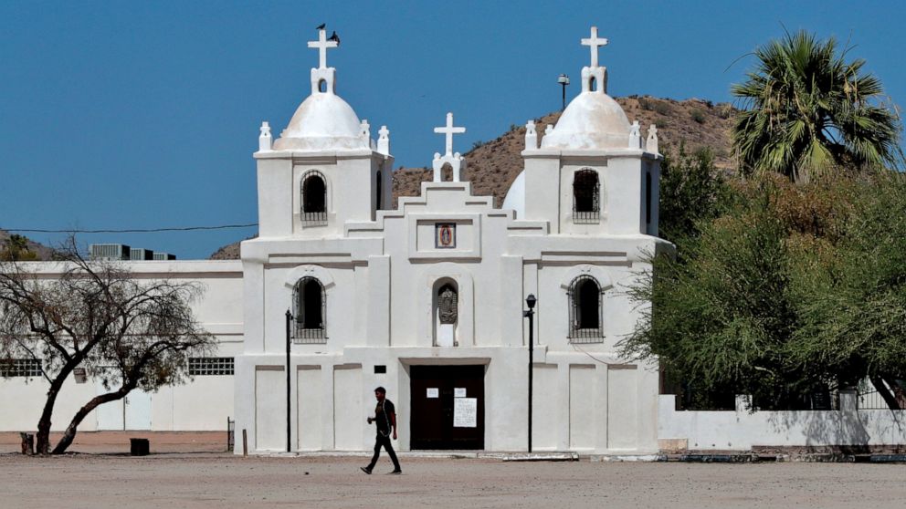 In this June 13, 2020, photo, a man walks past Our Lady of Guadalupe church in Guadalupe, Ariz. As the coronavirus spreads deeper across America, it's ravaging through the homes and communities of Latinos from the suburbs of the nation's capital to t