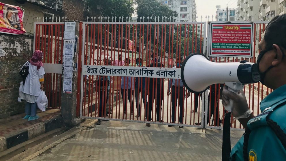 In this Monday, March 23, 3030 photo, a policeman urges residents not to come out of their homes as residents stand behind a gate, hours after the second death from COVID-19 was confirmed from the area, in Dhaka, Bangladesh. For most people, the new 