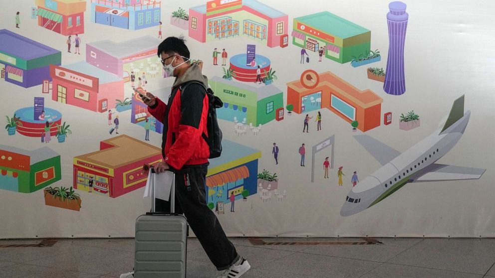 A traveller wearing a face mask with his luggage and passport walks by a poster depicting the Beijing airport on display at the international flight check in counter at the Beijing Capital International Airport in Beijing, Thursday, Dec. 29, 2022. Mo