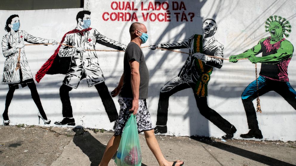 A man, wearing a protective face mask as a measure to curb the spread of the new coronavirus, walks past a mural depicting a tug-of-war between health workers and Brazil's President Jair Bolsonaro aided by a cartoon-styled coronavirus character, with