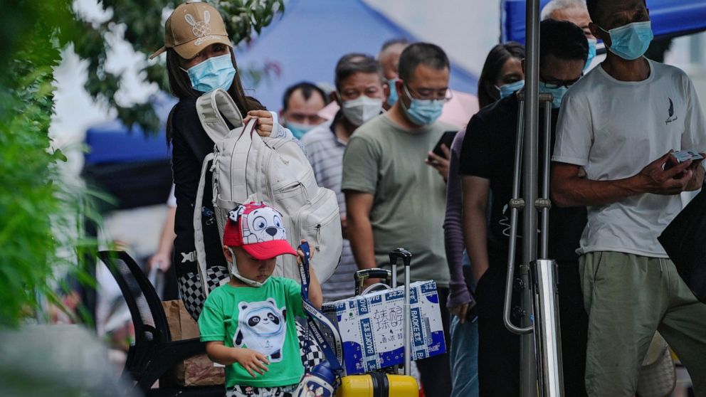 A woman and a child with their luggage stand watch as masked residents line up to get their routine COVID-19 throat swab at a coronavirus testing site in Beijing, Wednesday, Aug. 3, 2022. (AP Photo/Andy Wong)