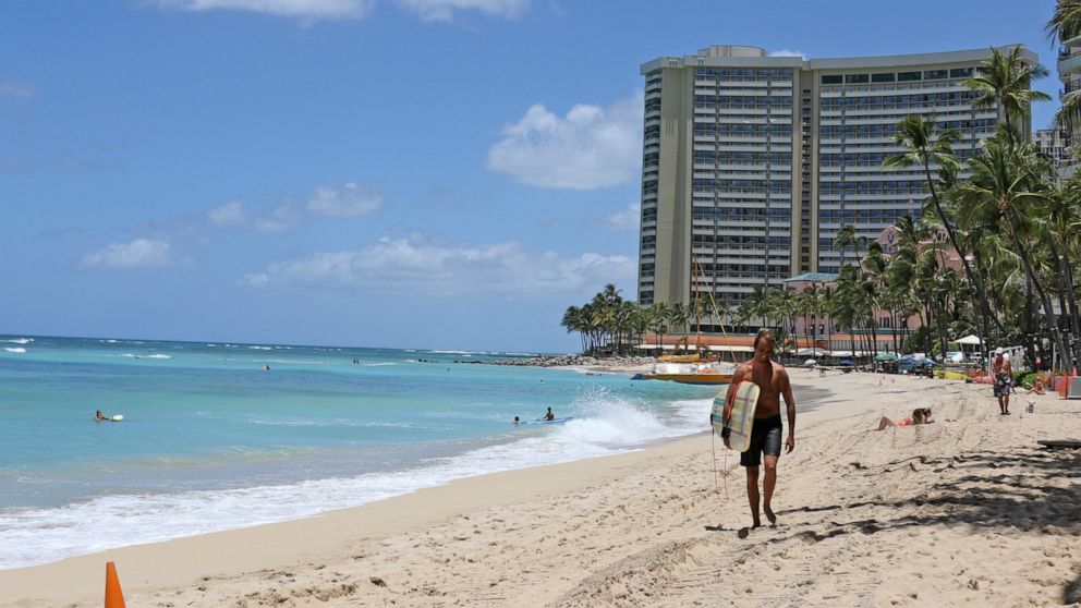 In this photo taken Friday, June 5, 2020, a surfer walks on a sparsely populated Waikiki beach in Honolulu. Hawaii faces unpleasant options for addressing a dramatic decline in tax revenues precipitated by the coronavirus pandemic and the shutdown of