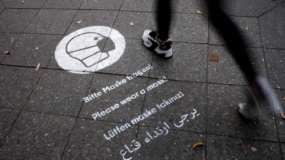 A pedestrian walks over a reminder to wear face masks sprayed on the sidewalk at the Karl-Marx-Strasse at the district Neukoelln in Berlin, Germany, Tuesday, Oct. 27, 2020. Karl-Marx-Strasse is one of the streets in the German capital where face mask