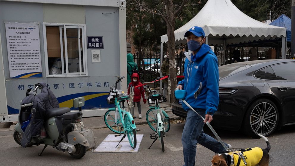 A man walks his dog past a COVID testing station in Beijing, Friday, Dec. 2, 2022. More cities eased restrictions, allowing shopping malls, supermarkets and other businesses to reopen following protests last weekend in Shanghai and other areas in whi