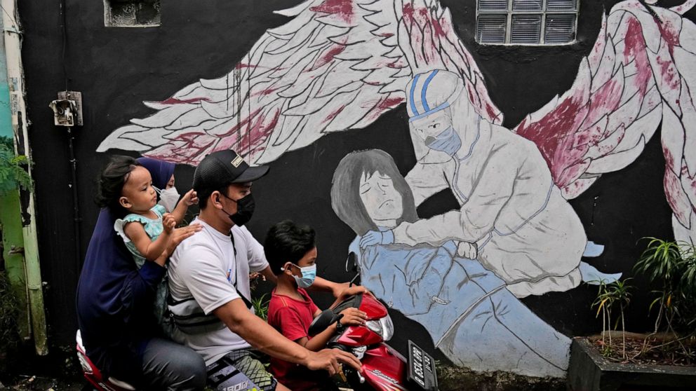 A family wearing masks to help curb the spread of coronavirus outbreak rides on a motorbike past a COVID-19-related mural honoring health workers in Jakarta, Indonesia, Saturday, Jan. 29, 2022. Indonesia is bracing for a third wave of COVID-19 infect