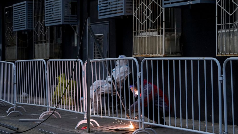 A worker in protective suit keeps watch as a worker welds on a steel frame to install metal barriers in a locked down neighborhood as part of COVID-19 controls in Beijing, Thursday, Nov. 24, 2022. China is expanding lockdowns, including in a central 