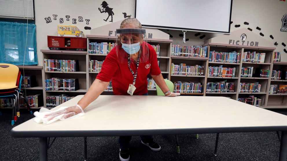 Wearing a mask and face guard as protection against the spread of COVID-19, Garland Independent School District custodian Camelia Tobon wipes down a table in the library at Stephens Elementary School in Rowlett, Texas, Wednesday, July 22, 2020.(AP Ph