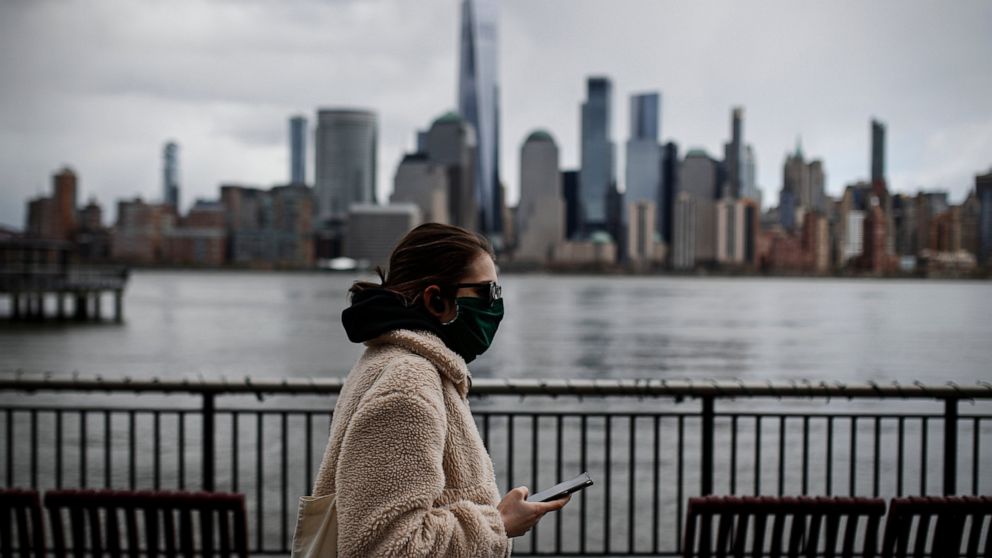 FILE- In this April 10, 2020 file photo a woman wearing a face mask sure to CVID-19 concerns walks along the Jersey City waterfront with the New York City skyline in the background. New York City may be the epicenter of the coronavirus outbreak but o