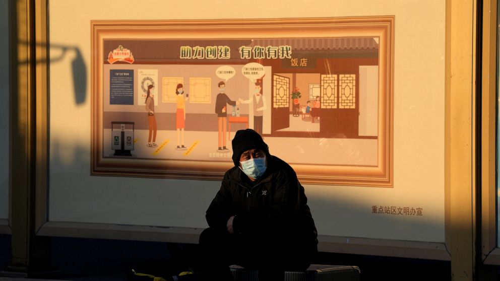 A man wearing a mask sits on his luggage near a mural depicting health checks before entry to a restaurant in Beijing, Thursday, Dec. 8, 2022. A day after China announced the rollback of some of its most stringent COVID-19 restrictions, people across
