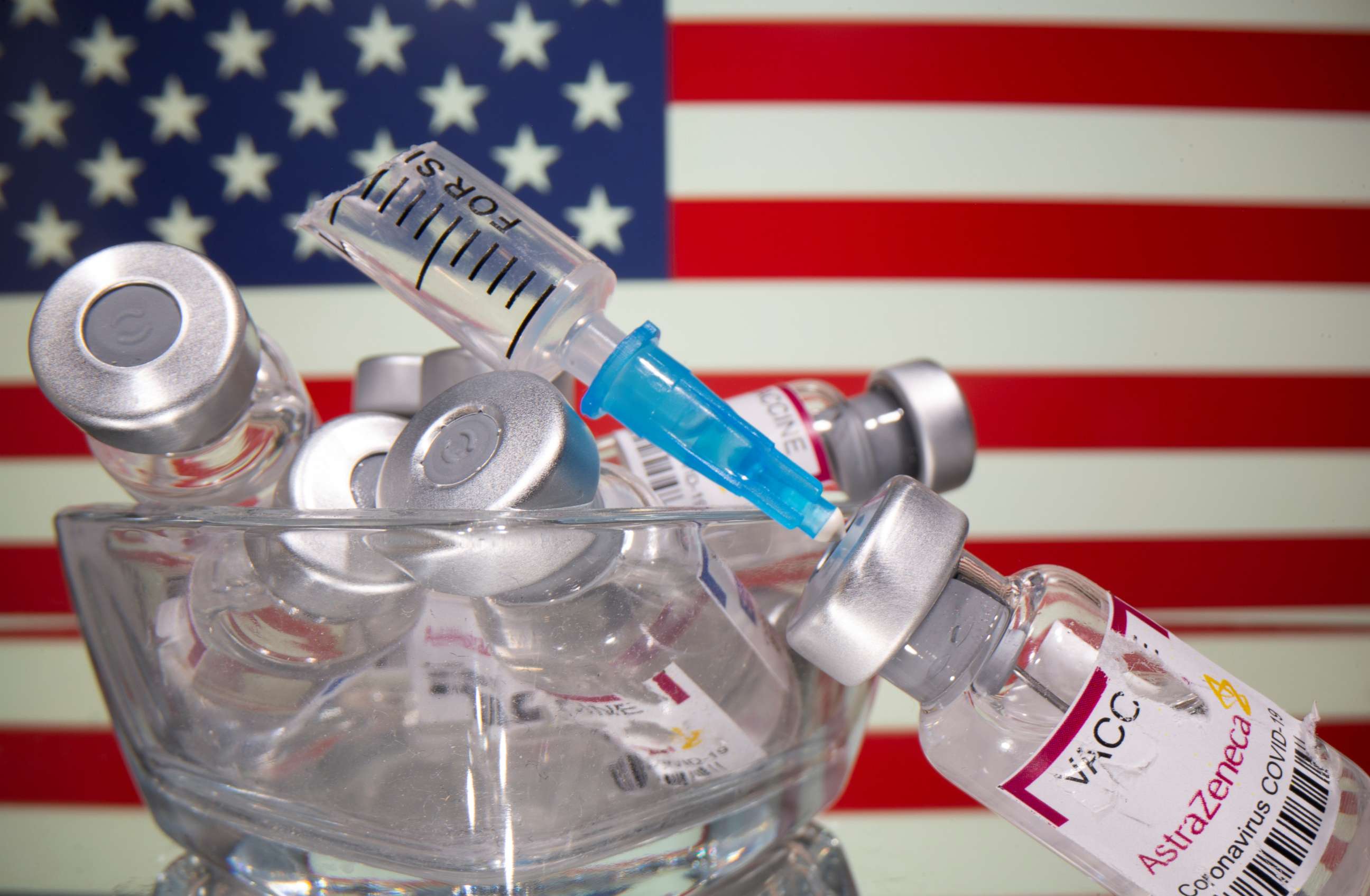 PHOTO: Vials labelled with broken sticker "AstraZeneca COVID-19 Coronavirus Vaccine" and a broken syringe are seen in front of a displayed U.S. flag in this illustration taken March 15, 2021.