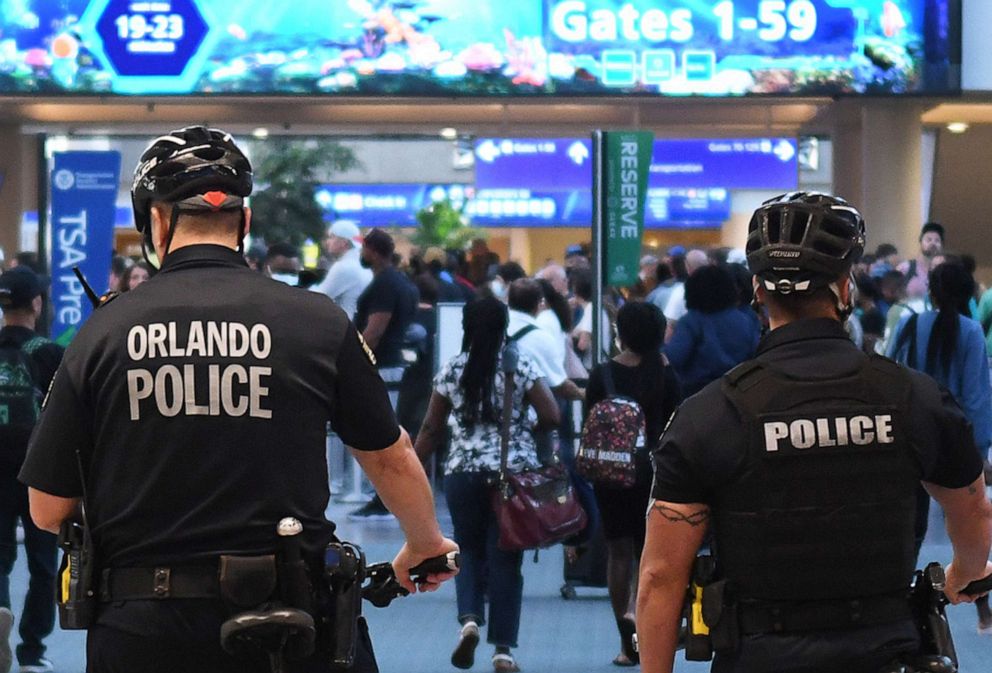 PHOTO: In this July 1, 2022, file photo, police officers watch as travelers make their way through a TSA screening line at Orlando International Airport ahead of the July 4 holiday. 