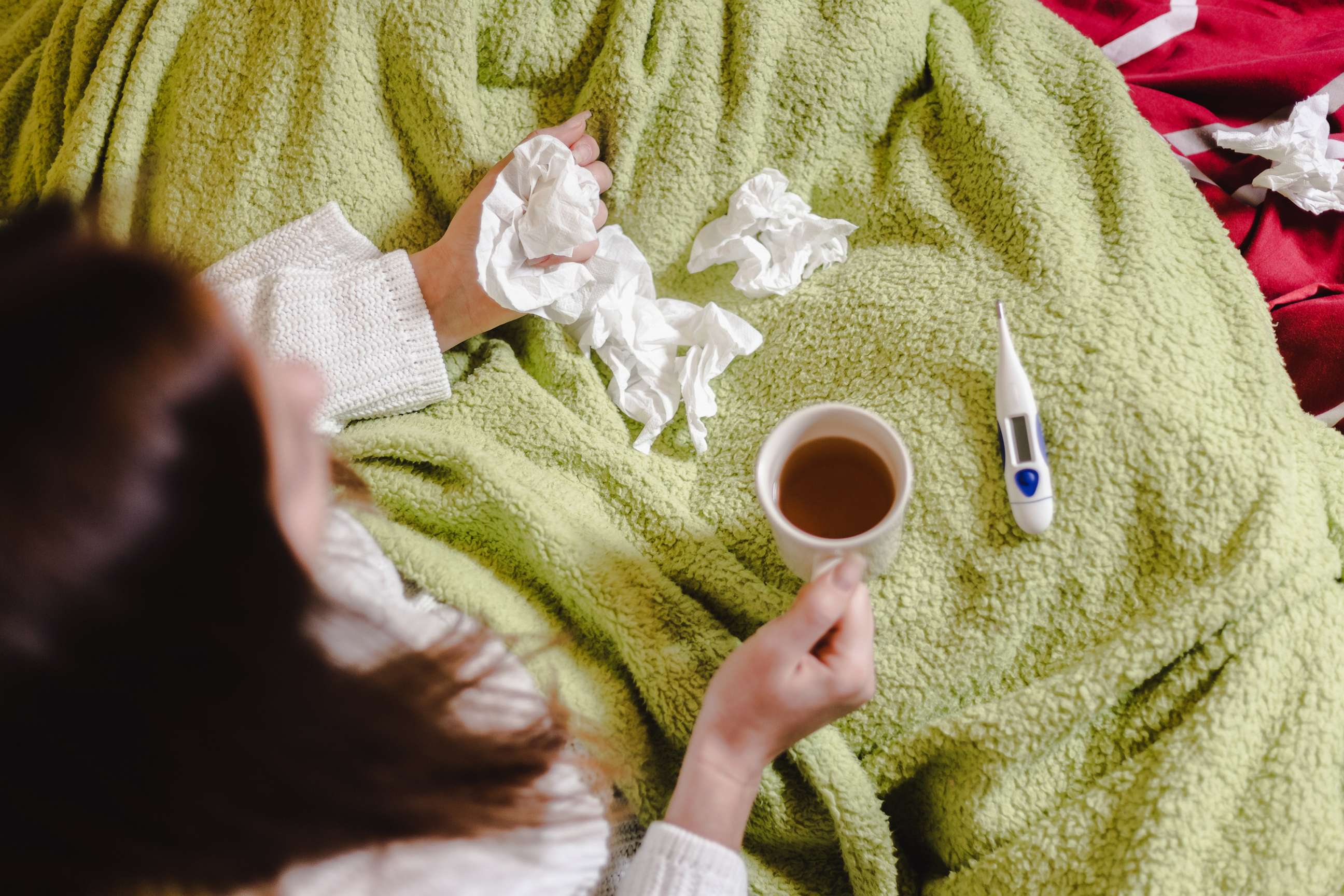PHOTO: A woman sits on her bed with a cold, surrounded by paper tissues, a cup of tea, and a thermometer.