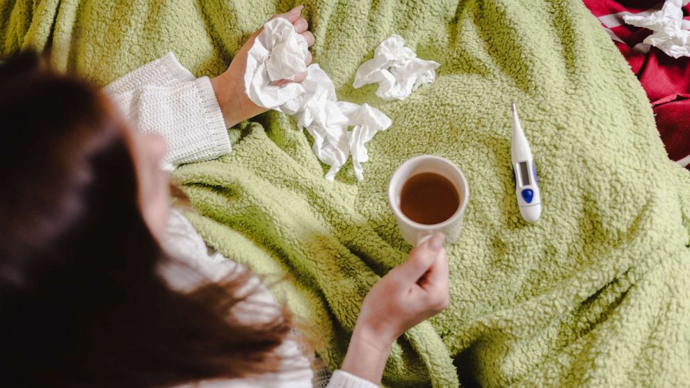 PHOTO: A woman sits on her bed with a cold, surrounded by paper tissues, a cup of tea, and a thermometer.