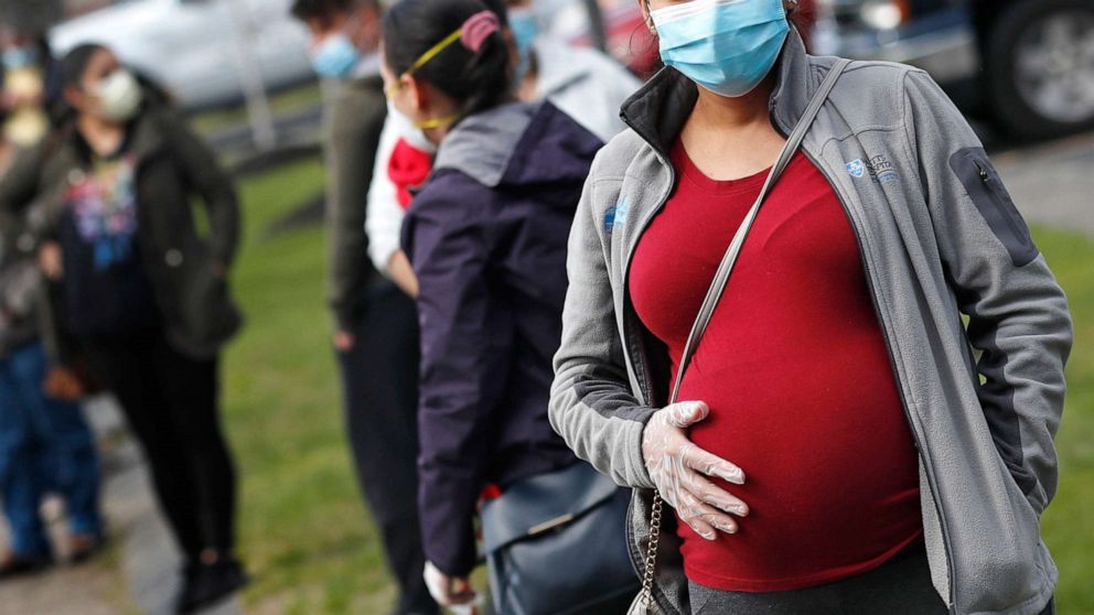 PHOTO: A pregnant woman wearing a face mask and gloves holds her belly as she waits in line for groceries at St. Mary's Church in Waltham, Mass, May 7, 2020.
