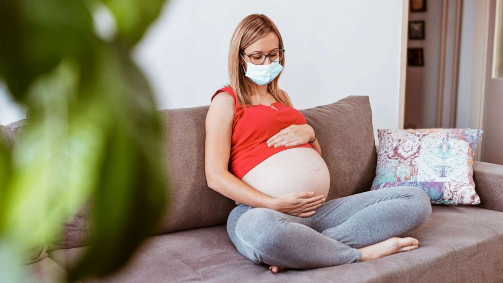 A pregnant woman wearing face mask is pictured seated in her living room in...
