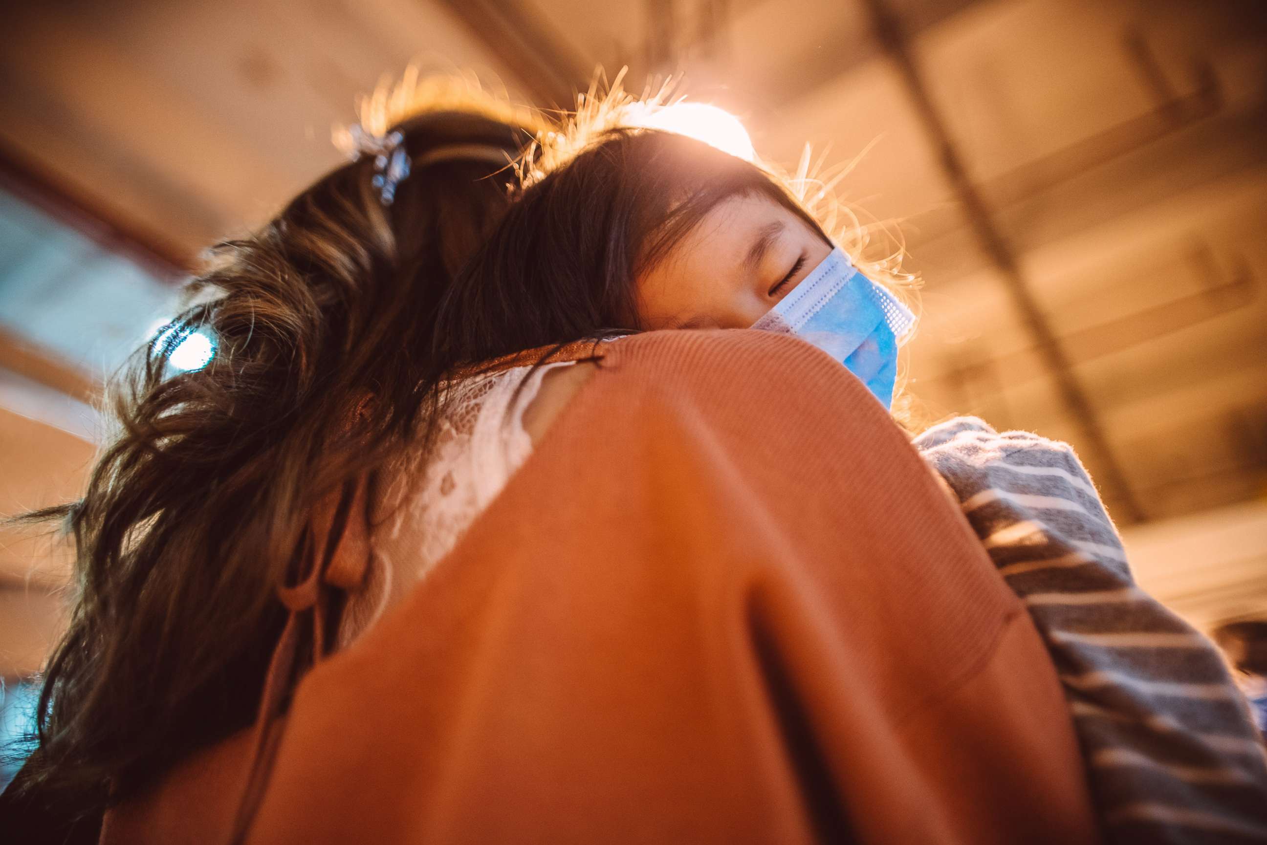 PHOTO: Back view of tired little girl in medical face mask falling asleep on mom's shoulder while strolling in street in the evening.