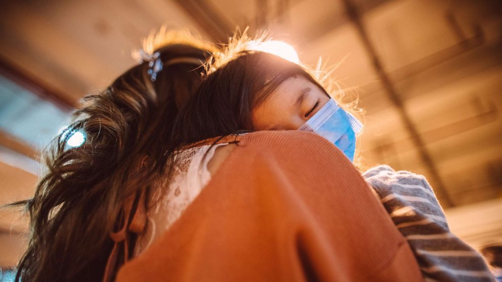 PHOTO: Back view of tired little girl in medical face mask falling asleep on mom's shoulder while strolling in street in the evening.